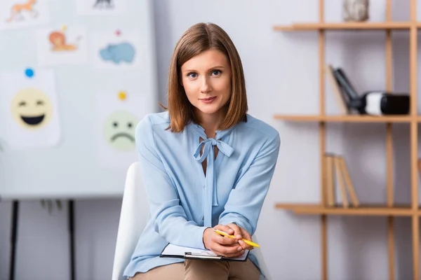 Psicóloga feminina positiva olhando para a câmera enquanto sentada na cadeira com escritório embaçado no fundo — Fotografia de Stock