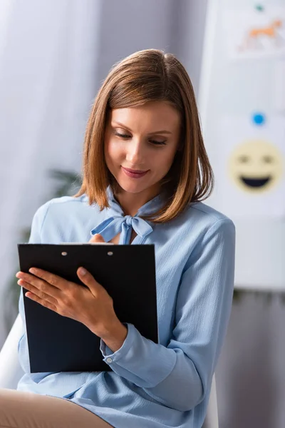 Sonriente psicóloga femenina escribiendo en portapapeles con un rotafolio borroso en el fondo - foto de stock
