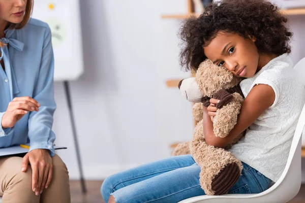 Deprimida chica afroamericana con autismo mirando a la cámara y abrazando oso de peluche cerca de psicólogo con oficina borrosa en el fondo - foto de stock