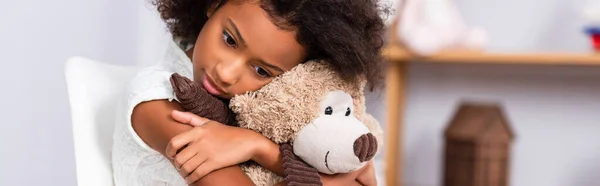 Deprimido afroamericano chica abrazando osito de peluche con oficina borrosa en el fondo, bandera - foto de stock