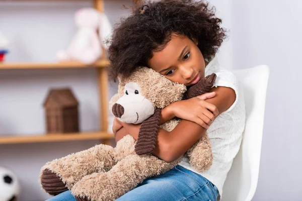 Deprimida chica afroamericana con autismo abrazando oso de peluche mientras está sentado en la silla con la oficina borrosa en el fondo - foto de stock