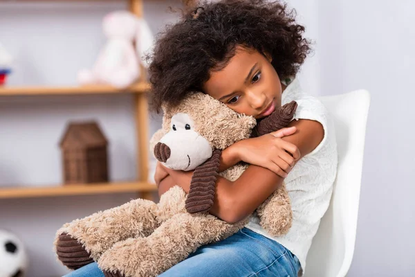 Déprimé afro-américaine fille avec autisme étreignant ours en peluche tout en étant assis sur une chaise avec un bureau flou sur fond — Photo de stock