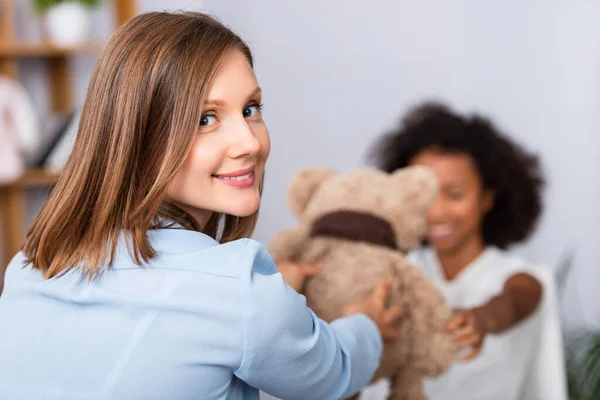 Glückliche Psychologin schaut in die Kamera, während sie einem afrikanisch-amerikanischen Mädchen einen Teddybär während einer Beratung über verschwommenen Hintergrund gibt — Stockfoto