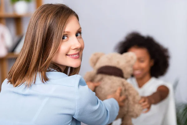 Psicólogo feliz olhando para a câmera ao dar ursinho de pelúcia para a menina americana africana durante a consulta sobre fundo borrado — Fotografia de Stock