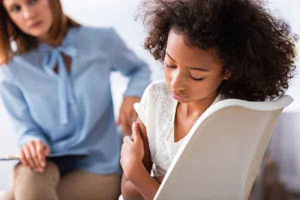 Upset african american girl with closed eyes sitting on chair during consultation with blurred psychologist on background — Stock Photo