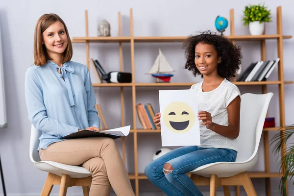 Sonriente chica afroamericana mostrando papel con expresión feliz mientras se sienta cerca de psicólogo sobre fondo borroso - foto de stock