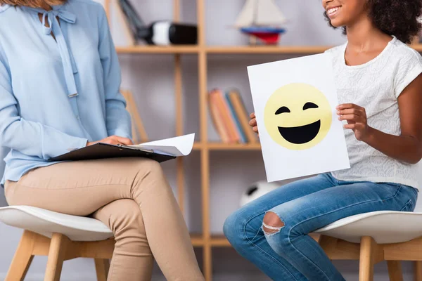 Vista recortada de chica afroamericana con expresión feliz en papel sentado cerca de psicólogo sobre fondo borroso - foto de stock