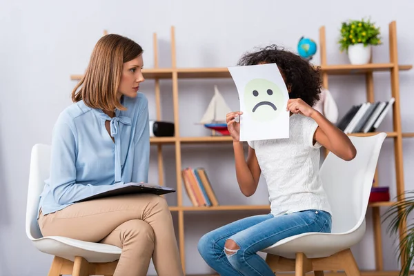 Psychologist looking at african american girl covering face with unhappy expression on paper with blurred office on background — Stock Photo