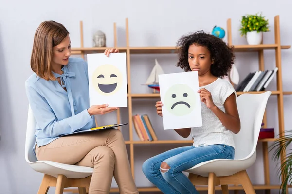 Psychologue et afro-américaine avec autisme montrant des expressions tristes et souriantes sur les papiers tout en étant assis sur des chaises pendant la consultation — Photo de stock