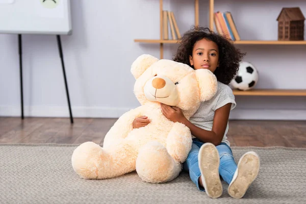 Fille afro-américaine bouleversée avec autisme regardant la caméra et étreignant ours en peluche tout en étant assis sur le sol avec un bureau flou sur fond — Photo de stock