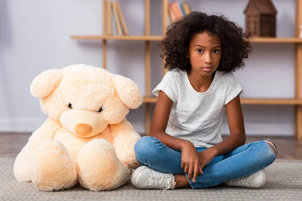 Deprimida chica afroamericana con las piernas cruzadas sentado en el suelo cerca de oso de peluche con oficina borrosa en el fondo - foto de stock