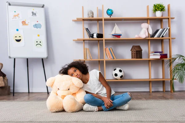 Deprimida afroamericana chica apoyada en osito de peluche mientras se sienta en el suelo cerca de estantes y rotafolio con imágenes en la oficina - foto de stock