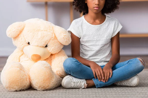 Vista recortada de chica afroamericana con piernas cruzadas sentada cerca de oso de peluche en el suelo sobre fondo borroso - foto de stock