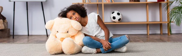 Deprimida chica afroamericana con autismo mirando hacia otro lado mientras se apoya en el oso de peluche en el suelo cerca de los estantes en la oficina, pancarta - foto de stock