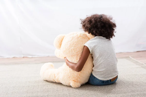 Vue arrière de fille afro-américaine solitaire avec autisme étreignant ours en peluche tout en étant assis sur le sol près du rideau — Photo de stock