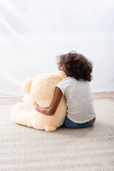 Vista trasera de la chica afroamericana solitaria abrazando oso de peluche mientras está sentado en el suelo cerca de la cortina - foto de stock