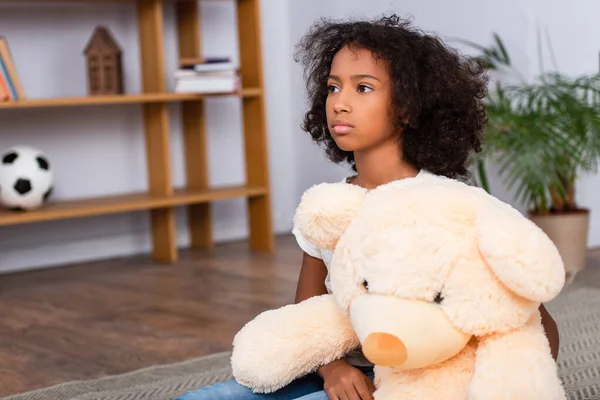 Upset african american girl looking away while hugging teddy bear on floor with blurred office on background — Stock Photo