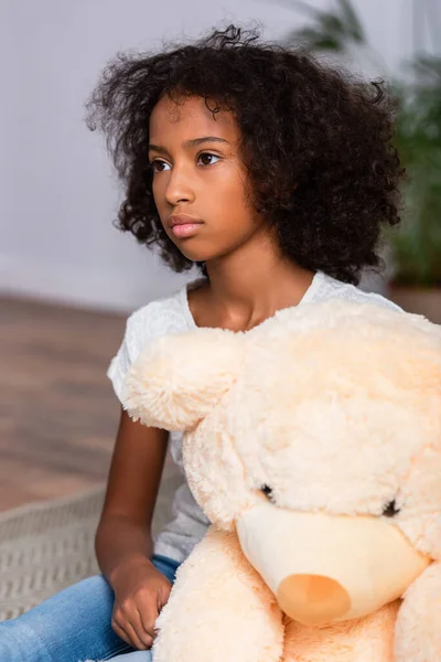 Niedergeschlagenes afrikanisch-amerikanisches Mädchen blickt in der Nähe von Teddybär auf dem Boden auf verschwommenem Hintergrund weg — Stockfoto