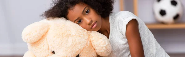 Menina afro-americana perturbada olhando para a câmera enquanto se inclina no ursinho de pelúcia no fundo borrado, banner — Fotografia de Stock