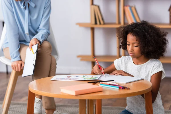 African american girl drawing with colored pencils on coffee table near psychologist with clipboard on blurred background — Stock Photo