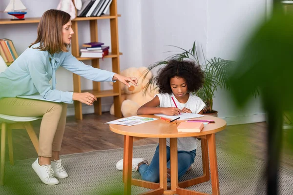 Pleine longueur de psychologue pointant du doigt l'album près d'une fille afro-américaine dessinant sur une table basse au premier plan flou — Photo de stock