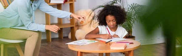 Psychologue pointant du doigt l'album près d'une fille afro-américaine dessinant avec des crayons de couleur au premier plan flou, bannière — Photo de stock