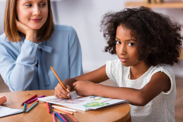 Menina afro-americana olhando para a câmera ao desenhar com lápis de cor perto de psicólogo positivo no fundo borrado — Fotografia de Stock