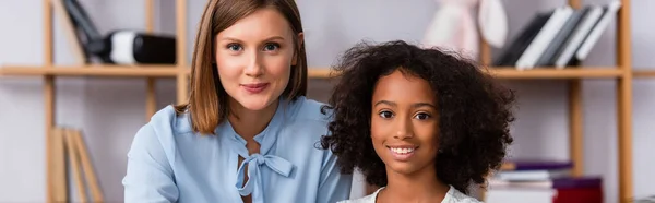 Happy multicultural psychologist and girl looking at camera on blurred background, banner — Stock Photo