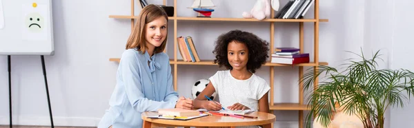 Happy psychologist and african american girl looking at camera near coffee table with colored pencils in office, banner — Stock Photo