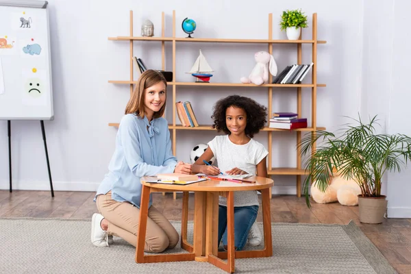 Longitud completa de psicólogo multicultural feliz y chica mirando a la cámara mientras está sentado cerca de la mesa de café en la oficina - foto de stock