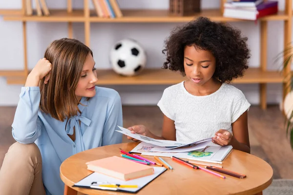 Positive psychologist looking at african american girl with pictures at coffee table on blurred background — Stock Photo