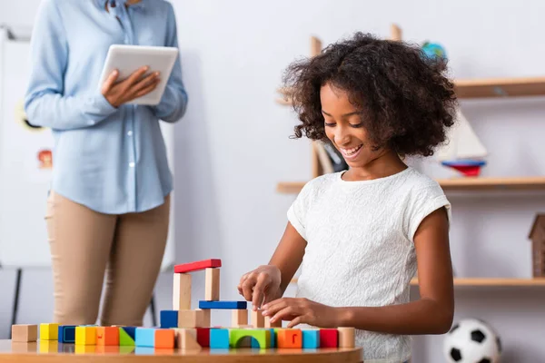 Happy african american girl playing with colorful wooden blocks at coffee table with blurred psychologist on background — Stock Photo