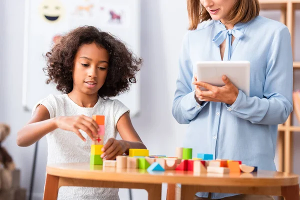 African american girl playing with wooden blocks near psychologist holding digital tablet with blurred coffee table on background — Stock Photo