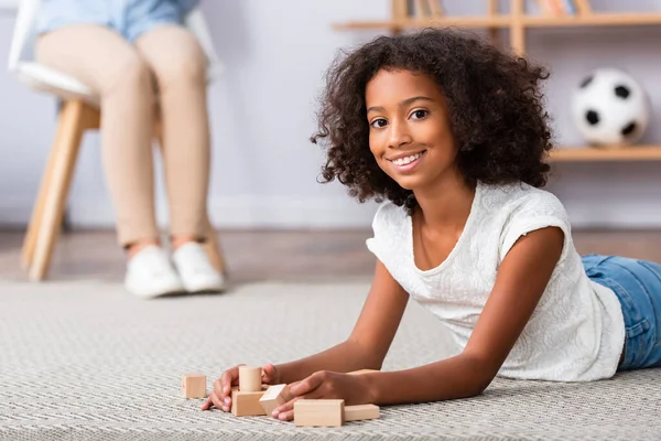 Souriant afro-américaine fille regardant caméra et jouer avec des blocs de bois tout en étant couché sur le sol sur fond flou — Photo de stock