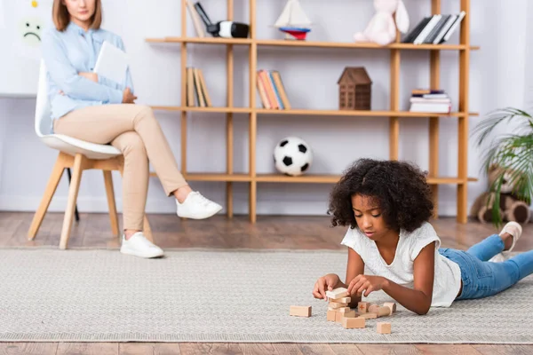 Focada menina afro-americana brincando com blocos de madeira enquanto deitado no chão com escritório borrado no fundo — Fotografia de Stock