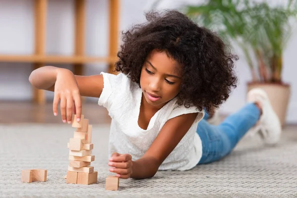 Concentrato afroamericano ragazza giocare con blocchi di legno mentre sdraiato sul pavimento con ufficio offuscata su sfondo — Foto stock