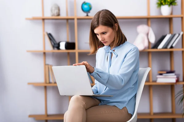 Konzentrierter Psychologe mit Laptop, während er im Büro auf einem Stuhl vor verschwommenem Hintergrund sitzt — Stockfoto