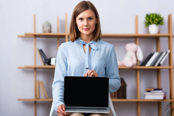 Positive psychologist looking at camera while showing laptop with blank screen on blurred background — Stock Photo