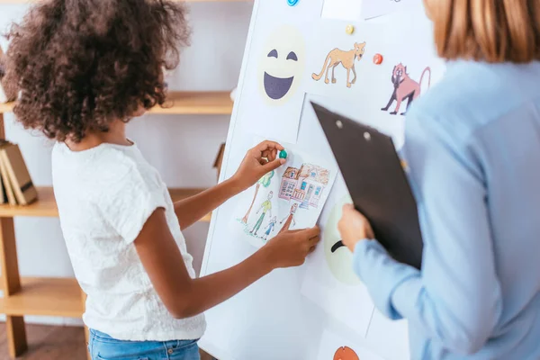 Africano menina americana pendurado desenho no quadro branco perto de imagens, enquanto está perto psicólogo em primeiro plano borrado — Fotografia de Stock