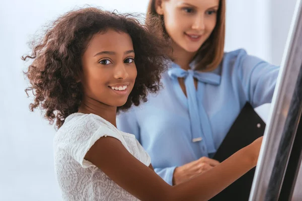 Happy african american girl looking at camera near whiteboard with blurred psychologist on background — Stock Photo
