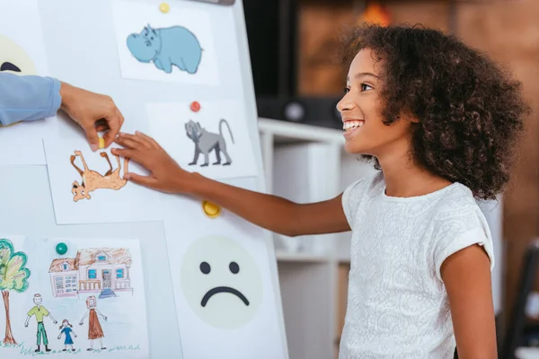 Menina americana africana alegre tocando imagem no quadro branco perto da mão do psicólogo no fundo borrado — Fotografia de Stock