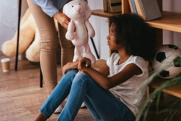 Fille afro-américaine bouleversée avec autisme regardant un jouet doux dans les mains d'un psychologue se tenant près sur le premier plan flou — Photo de stock