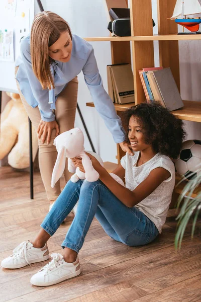 Psicólogo inclinado hacia adelante a la chica afroamericana feliz con juguete suave sentado en el suelo en primer plano borroso - foto de stock