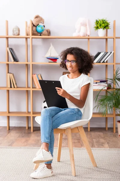 Die ganze Länge des lächelnden afrikanisch-amerikanischen Mädchens mit Brille und Klemmbrett, das im Büro auf einem Stuhl sitzt und wegschaut — Stockfoto