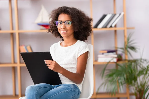 Lächelndes afrikanisch-amerikanisches Mädchen mit Brille und Klemmbrett, das auf einem Stuhl vor verschwommenem Hintergrund in die Kamera blickt — Stockfoto