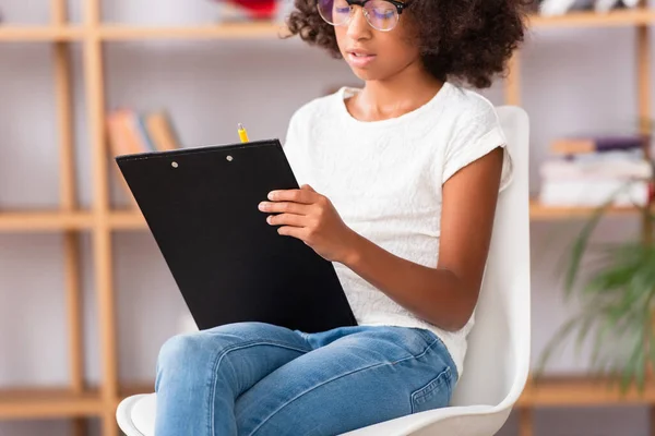 Vue recadrée d'une fille afro-américaine écrivant sur un presse-papiers assis sur une chaise sur un fond flou — Photo de stock