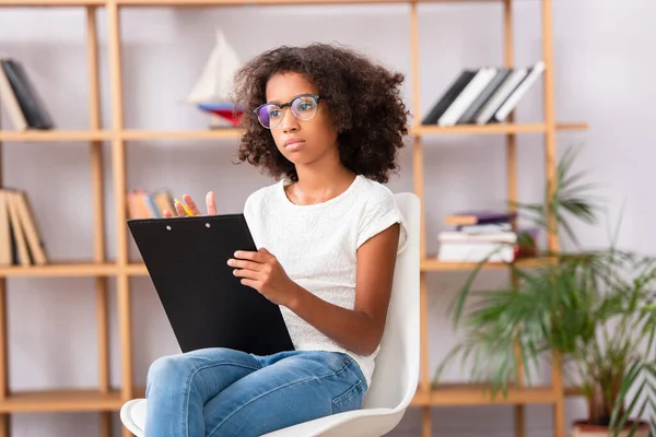 African american girl in eyeglasses with clipboard looking away while sitting on chair on blurred background — Stock Photo