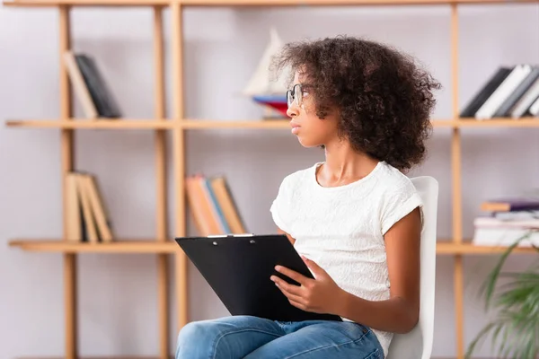 African american girl in eyeglasses with clipboard looking away while sitting on chair on blurred background — Stock Photo