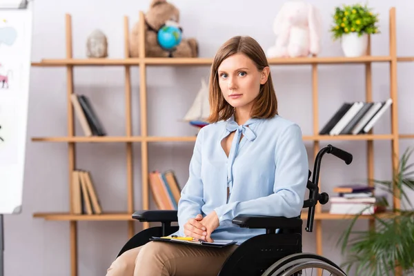 Disabled psychologist with clipboard looking at camera while sitting in wheelchair on blurred background — Stock Photo