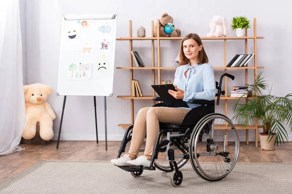 Full length of positive psychologist with clipboard looking at camera while sitting in wheelchair in office — Stock Photo